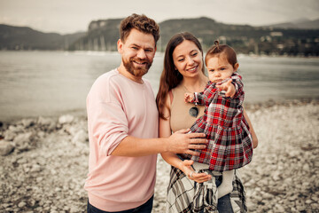 Young happy family spending their vacation on Italian Garda lake coast, bearded daddy, smiling mom, their little toddler girl in red checkered dress pointing with finger