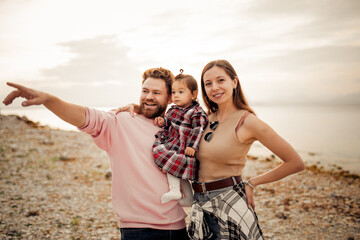 Young happy family spending their vacation on Italian Garda lake coast, bearded daddy pointing with finger, smiling mom, their little toddler girl in red checkered dress 