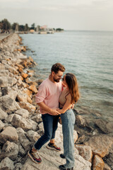 Fototapeta premium Young attractive cute couple in romantic honeymoon standing at Garda lake stones kissing and embracing each other at the sunset, bearded curly man and brunette girl in sunglasses 