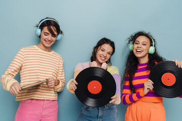 Three beautiful girls in headphones holding vinyl records and smiling isolated over blue background
