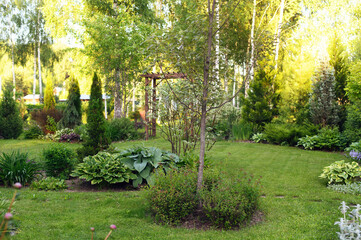 summer private Europe garden view with wooden archway, hostas, conifers and shrubs. Lawn with curvy...