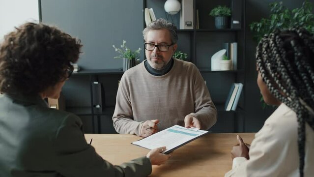 Medium shot of front view of grey-haired Caucasian man sitting in front of black female HR who talking to him smiling, with Caucasian woman sitting beside her, listening. Man amiably smiling back
