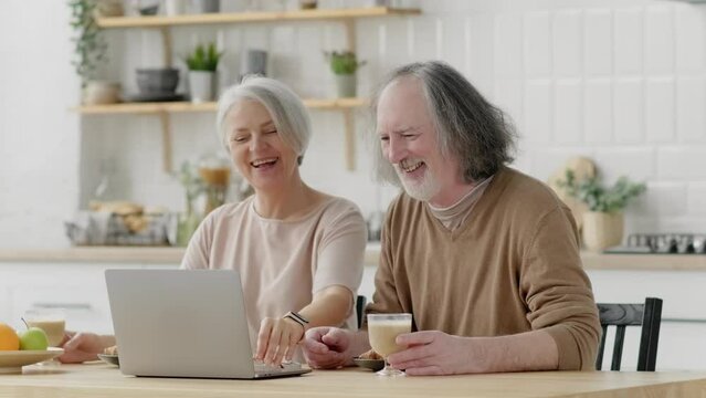 Happy middle aged 60s family couple having fun using laptop computer at home. Smiling senior mature husband and wife laughing watching funny content or having virtual meeting in living room.