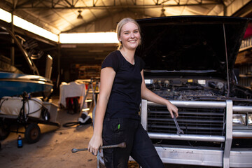 Young female aussie mechanic holding tools and leaning on a car in need of repair in workshop garage