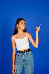 Fototapeta na wymiar Portrait of pretty teen girl in casual clothes pointing index finger up on empty blue background, looking up away. Cute teenage lady expressing. Advertising presentation concept. Copy ad text space