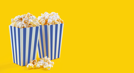 Two blue white striped carton buckets with tasty cheese popcorn, isolated on yellow background. Box with scattering of popcorn grains. Fast food, movies, cinema and entertainment concept.