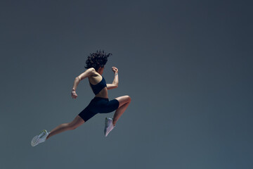 Athletic art. Dynamic image of sportive woman in motion, jumping, running, training against grey...
