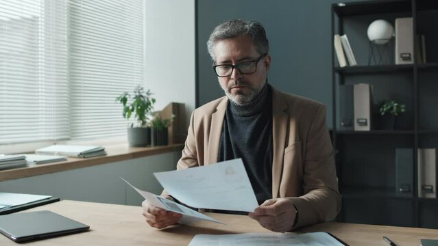 Medium shot of senior male HR looking through CVs, sitting in light office beside window, then seriously looking at camera, front viewMedium shot of senior male HR looking through CVs, sitting in ligh