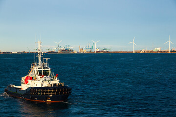 A tug boat sailing in the bay of the seaport of Fos-sur-Mer, France, with cranes and wind...