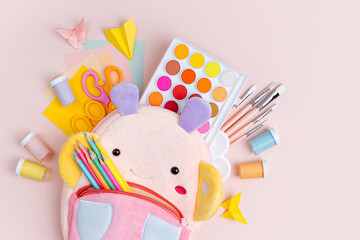 Opened kids backpack with stationery and supplies for drawing and craft on pink background. Various...