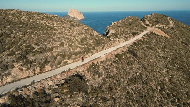 Aerial video of female cyclists in pink jersey descending on rural road on the hill with a sea view and Penon de Ifach rock in the background. Cycling in Calpe, Costa Blanca concept video.