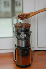 The roasted coffee beans are poured from a wooden spoon into an electric coffee grinder.