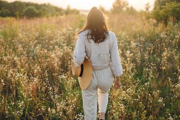 Stylish boho woman with straw hat walking among wildflowers in warm sunset light. Atmospheric moment. Summer delight and travel. Young female in rustic linen cloth relaxing in summer meadow. - obrazy, fototapety, plakaty