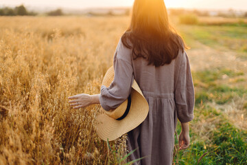 Stylish woman with straw hat walking at oat field in sunset light. Atmospheric tranquil moment....