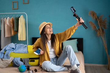 Social media vlogging concept. Young travel vlogger recording video content on smartphone, sitting on bed near the suitcase, speaking to her blog about journey.