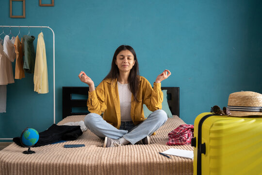 Young traveler woman meditating sitting near the suitcase on the bed at home, hold hands in yoga gesture. Female passenger traveling abroad on weekend. Air flight journey.