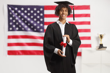 African american male student in a graduation gown holding a diploma and standing in front of USA...
