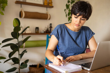 Young latin woman with a short hair taking notes from laptop at the home office. She is sitting in the apartment full of plants.