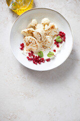 Plate with roasted cauliflower florets, cream-cheese and pomegranate seeds, flat lay on a light-beige stone background, vertical shot, copy space