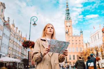 Attractive lost young female tourist holding a paper map on central square in Gdansk old town. Traveling Europe in autumn. High quality photo