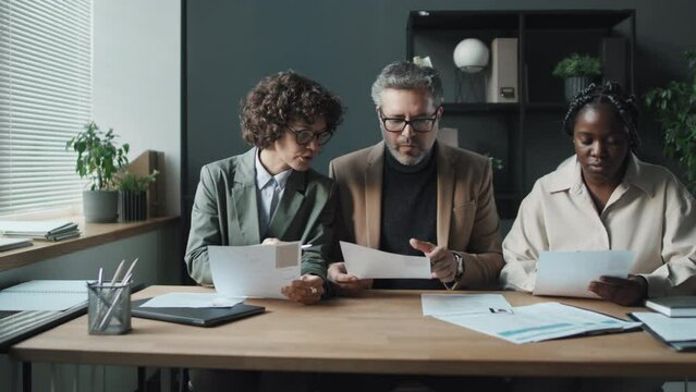 Medium shot of three HRs, Caucasian woman, senior Caucasian man, young black woman sitting in office, slowly looking through CVs, then raising their heads and smiling at camera, front view