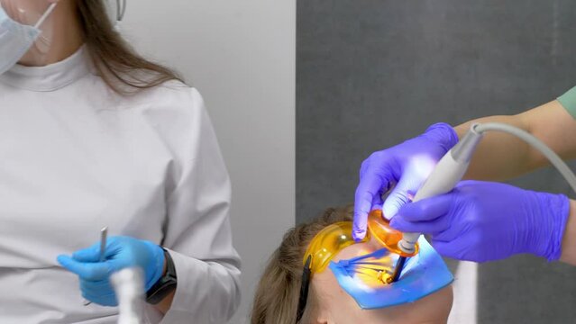 Tooth restoration with filling and polymerization lamp. Young girl during dental filling drying procedure with ultraviolet light curing in dental clinic. Work of doctor and assistant.