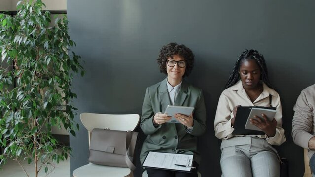 Medium shot of Caucasian woman, black woman and Caucasian man waiting in line for job interview, looking through their CVs, then Caucasian woman taking eyes from her CV, looking at camera and smiling