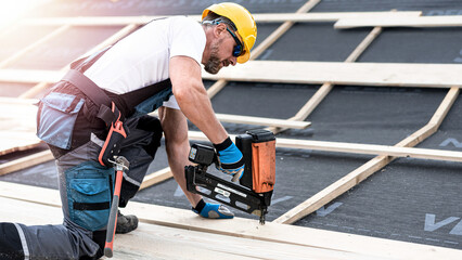 The carpenter nails a timber board using an electric nailer while working on a roof. - obrazy, fototapety, plakaty