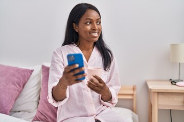 Fototapeta premium Young african american woman drinking coffee using smartphone at bedroom