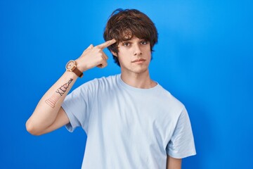 Naklejka premium Hispanic young man standing over blue background pointing unhappy to pimple on forehead, ugly infection of blackhead. acne and skin problem