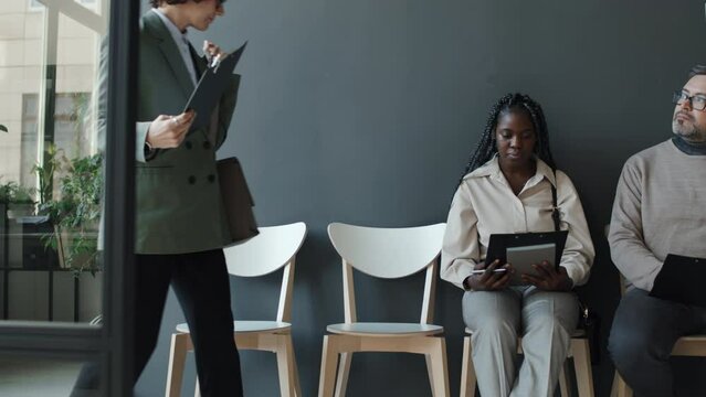Young Caucasian woman entering hall and sitting beside black woman and Caucasian man, smiling to them, all holding their CVs, waiting for interview, black woman taking her eyes off and looking to side