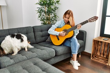 Young caucasian woman playing classical guitar sitting on sofa with dog at home