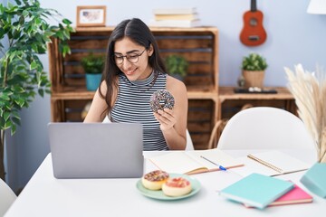 Obraz na płótnie Canvas Young hispanic girl eating doughnut studying at home