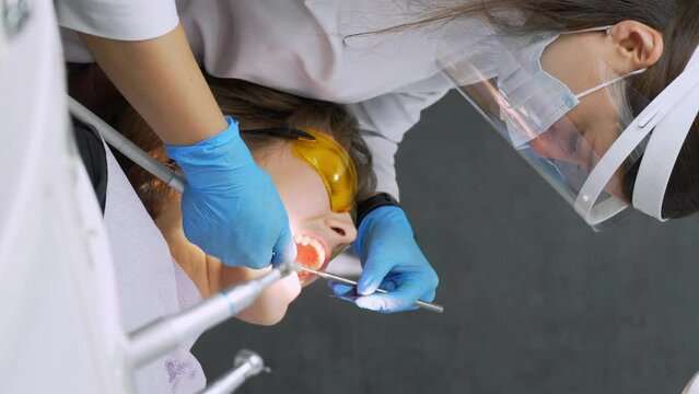Female dentist and assistant removing dental calculus from teeth. Visit is in proffessional dental clinic. Woman sits on dental chair. Drilling and treatment of tooth, filling. vertical video