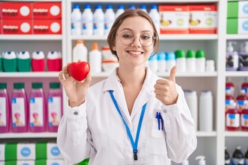 Young caucasian woman working at pharmacy drugstore holding red heart smiling happy and positive,...