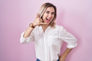 Young beautiful woman standing over pink background smiling doing phone gesture with hand and fingers like talking on the telephone. communicating concepts.