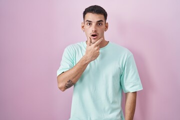 Handsome hispanic man standing over pink background looking fascinated with disbelief, surprise and amazed expression with hands on chin