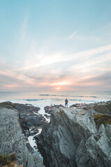 Fototapeta premium Fearless man with a backpack watching the sunset over the Atlantic Ocean in Porto Covo, Portugal. A hiker stands on the edge of a cliff looking into his future. Self-awareness