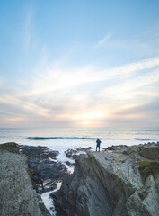 Fearless man with a backpack watching the sunset over the Atlantic Ocean in Porto Covo, Portugal. A hiker stands on the edge of a cliff looking into his future. Self-awareness