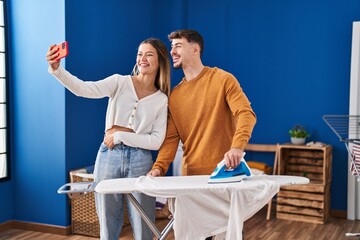 Young man and woman couple ironing clothes make selfie by the smartphone at laundry room