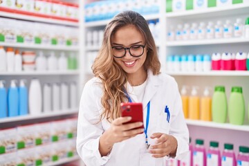 Young beautiful hispanic woman pharmacist using smartphone working at pharmacy