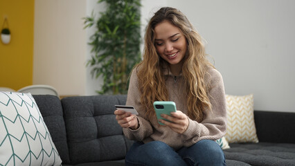 Young beautiful hispanic woman using smartphone and credit card sitting on sofa at home