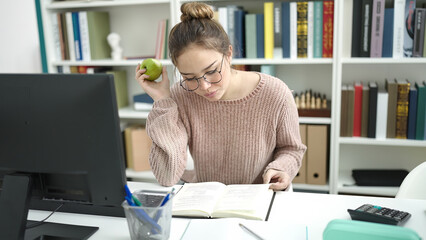 Young beautiful hispanic woman student reading book eating apple at library university