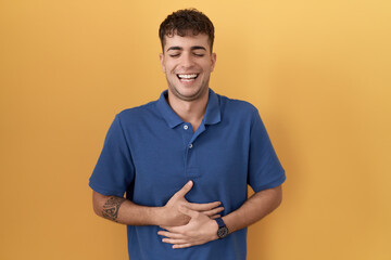 Young hispanic man standing over yellow background smiling and laughing hard out loud because funny...