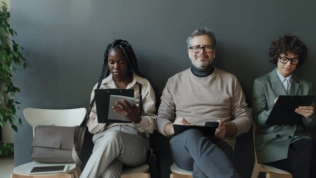 Medium shot of Caucasian woman, black woman and Caucasian man waiting in line for interview, looking through their CVs, then Caucasian man taking eyes from his CV, amiably looking at camera, smiling