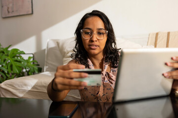 Latin woman using credit card at home. She is using digital tablet to check her bank statement...