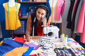 Fototapeta premium Young beautiful hispanic woman tailor talking on smartphone writing on notebook at clothing factory