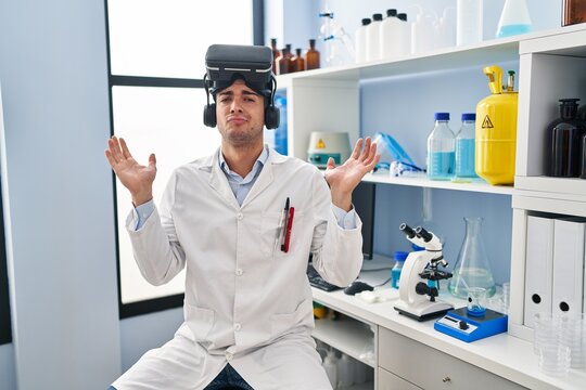 Young hispanic man working at scientist laboratory wearing vr glasses shouting and screaming loud to side with hand on mouth. communication concept.