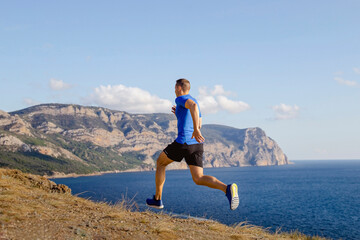 lean male runner run mountain trail along seashore, sports clothes blue shirt and black shorts, summer jogging along coast