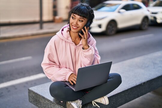 Young woman using laptop talking on the smartphone sitting on bench at street